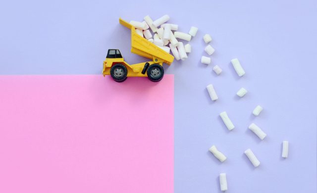 Yellow little toy dump truck throws marshmallow pieces from its raised back on a pastel violet and pink background. Flat lay minimal top view.