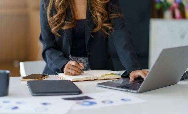 Close-up of a businesswoman sitting at a desk concentrating on fluently inspecting work using his laptop and several documents on the table at the office