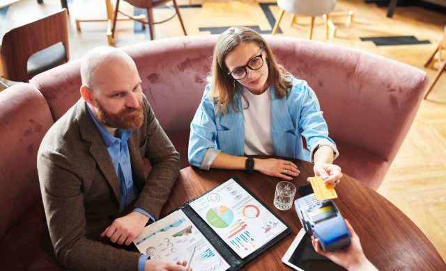 Above view of business people sitting at table with financial report and paying with smartphone in restaurant