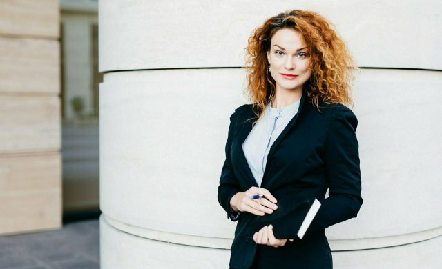 Pleasant-looking curly woman dressed formally, holding notebook in hands, going to have meeting wit her business partners. Gorgeous business lady writing in diary book. Professionalism concept
