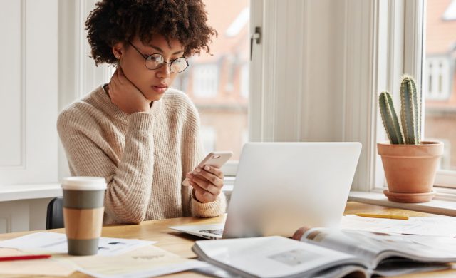 Serious female worker sits at desktop with laptop computer, surrounded with papers and journals, types sms message on smart phone device, connected to wireless internet, poses near window indoor